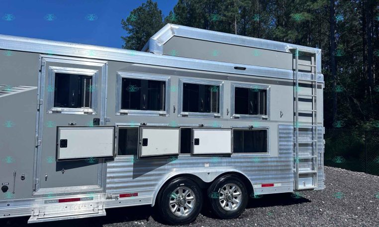 Silver aluminum horse trailer with three side windows, a rear ladder, and chrome trim parked on gravel with trees in the background.