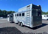 Silver aluminum horse trailer with side windows and a rear ramp parked on a gravel lot, trees in the background on a sunny day.