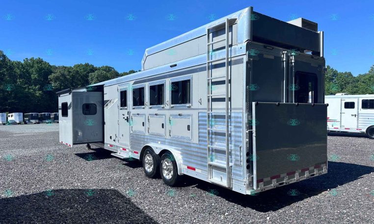 Silver aluminum horse trailer with side windows and a rear ramp parked on a gravel lot, trees in the background on a sunny day.