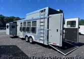 Silver horse trailer with ramp down on a gravel lot under a blue sky, side windows with bars, ladder attached to the front.