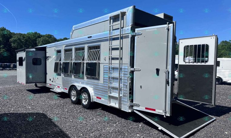 Silver horse trailer with ramp down on a gravel lot under a blue sky, side windows with bars, ladder attached to the front.