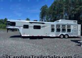 Silver multi-section horse trailer with windows and bars, parked on a gravel lot with trees in the background