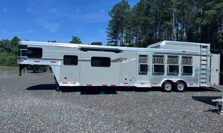 Silver multi-section horse trailer with windows and bars, parked on a gravel lot with trees in the background