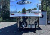 Rear view of a chrome SMC Trailers trailer parked on gravel, with a logo and reflective surface against a forest backdrop.