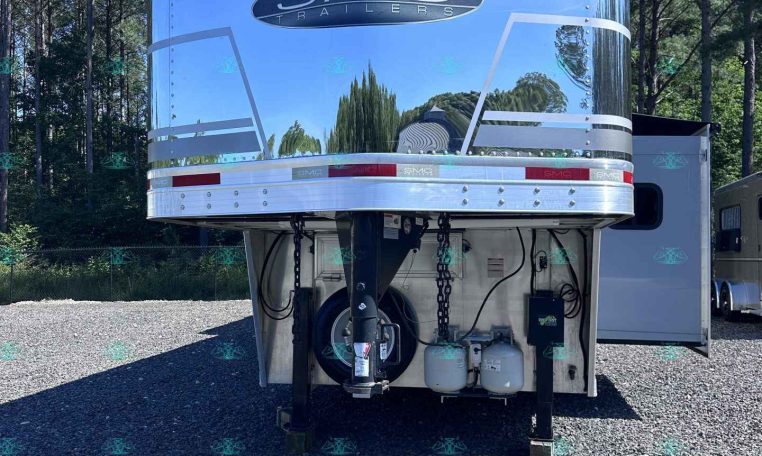 Rear view of a chrome SMC Trailers trailer parked on gravel, with a logo and reflective surface against a forest backdrop.