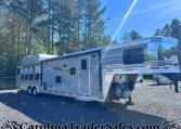 Silver fifth-wheel trailer parked on a gravel lot with tall pine trees and a bright blue sky in the background.