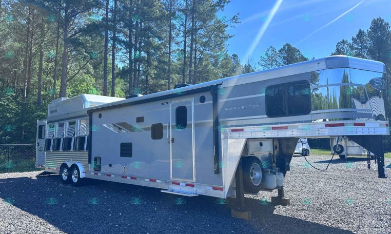 Silver fifth-wheel trailer parked on a gravel lot with tall pine trees and a bright blue sky in the background.