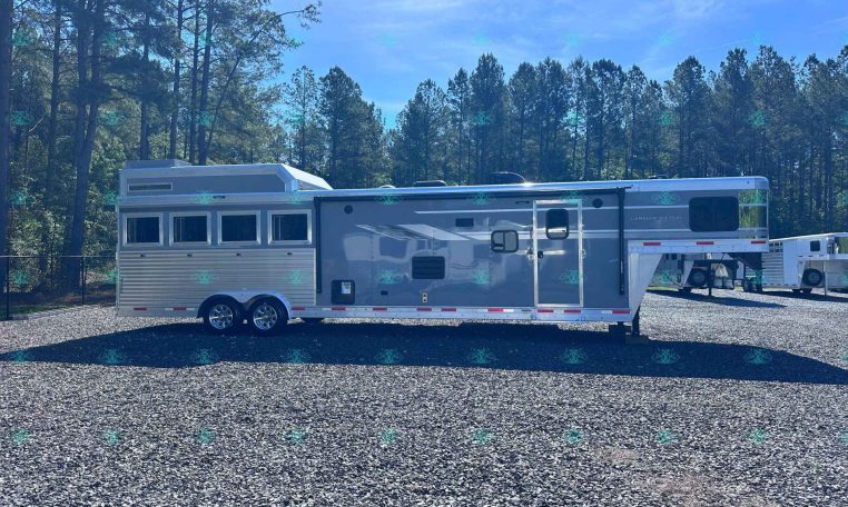 Large gray travel trailer parked on a gravel lot with a forest of tall pines in the background, under a clear blue sky.
