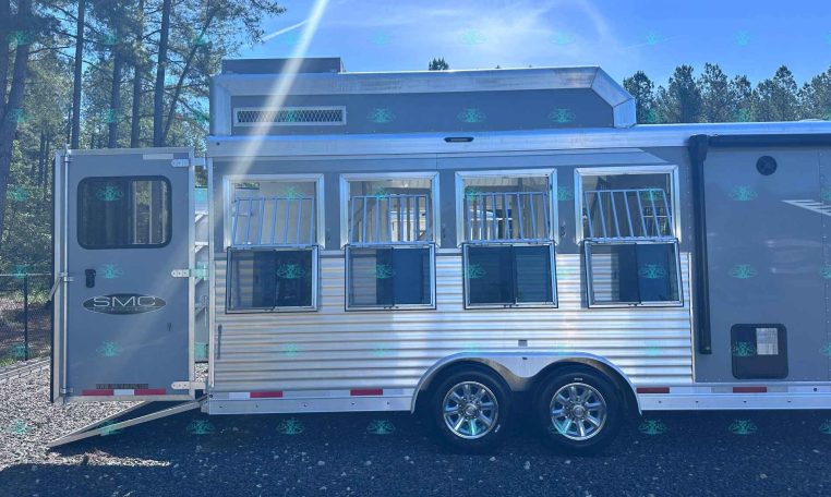 Silver horse trailer with open side windows and ramp down, parked on gravel with trees and blue sky in background; shows 'SMC Trailers' branding and website at bottom.