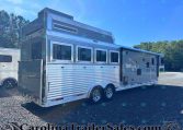 Silver livestock/horse trailer with multiple side windows and dual axles parked on gravel, trees in the background.