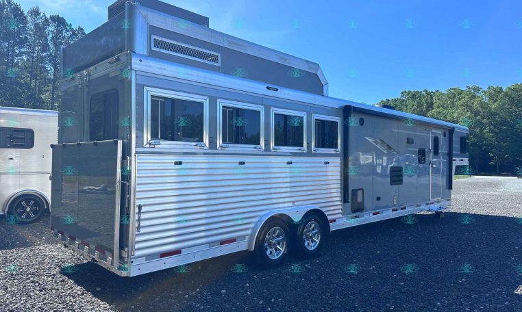 Silver livestock/horse trailer with multiple side windows and dual axles parked on gravel, trees in the background.