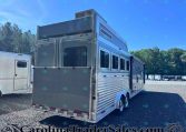 Silver aluminum horse trailer parked in a dealership lot with windows and rear doors visible, other trailers in the background on a sunny day.