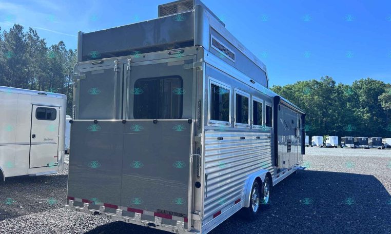 Silver aluminum horse trailer parked in a dealership lot with windows and rear doors visible, other trailers in the background on a sunny day.
