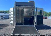 Open silver livestock trailer with doors swung wide and ramp down, parked on a gravel lot under a clear blue sky.