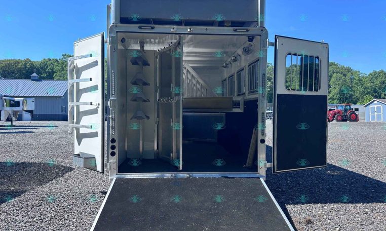 Open silver livestock trailer with doors swung wide and ramp down, parked on a gravel lot under a clear blue sky.