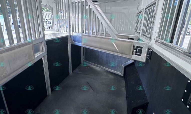 Interior of a metallic horse trailer with divided stalls and a slanted loading ramp, windows along the side, bright overhead lights.