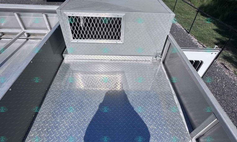 View from the back of a diamond-plate metal trailer looking toward a white horse trailer in a fenced, gravel lot with trees in the background.