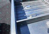 Close-up of a silver steel trailer bed with diamond-plate flooring and horizontal side rails; a person holds the edge in the lower left corner.