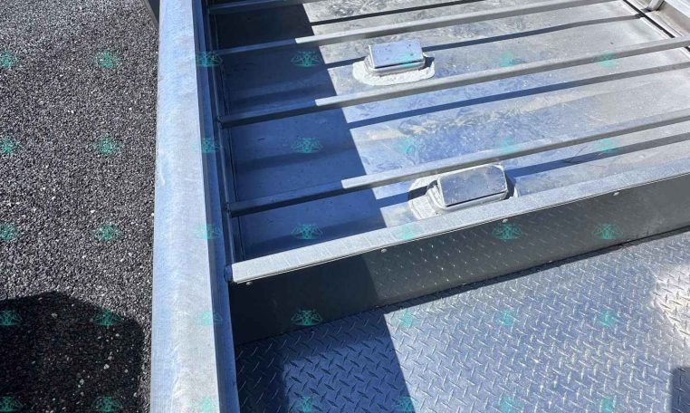 Close-up of a silver steel trailer bed with diamond-plate flooring and horizontal side rails; a person holds the edge in the lower left corner.