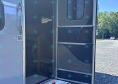 Open rear of a silver trailer with a doorway and small interior stairs visible inside, parked on a gravel lot under a blue sky.