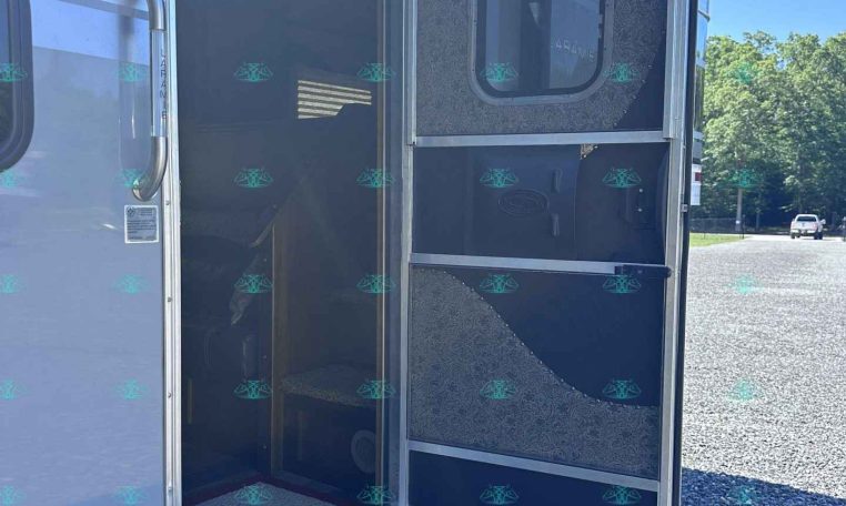 Open rear of a silver trailer with a doorway and small interior stairs visible inside, parked on a gravel lot under a blue sky.
