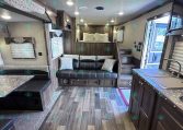 Interior of a modern RV living area with a gray sectional sofa, pillows, wood cabinetry, and a kitchen counter nearby.