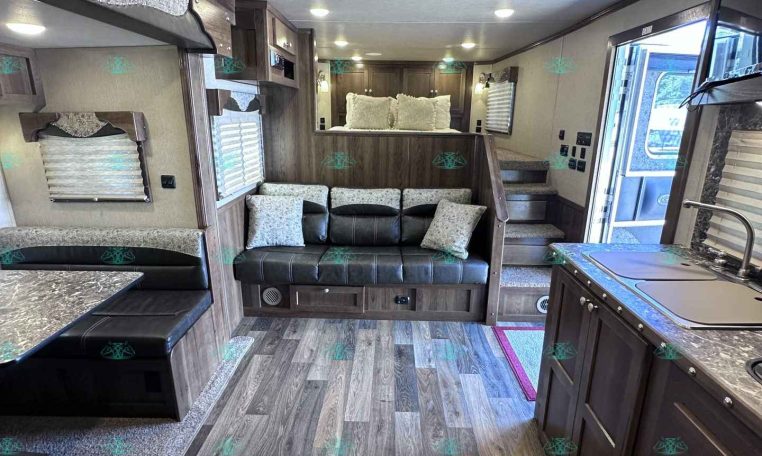 Interior of a modern RV living area with a gray sectional sofa, pillows, wood cabinetry, and a kitchen counter nearby.