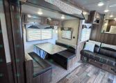 Interior of a recreational vehicle showing a booth-style dinette with a metal tabletop and dark leather seating, wood cabinets, and windows with blinds.