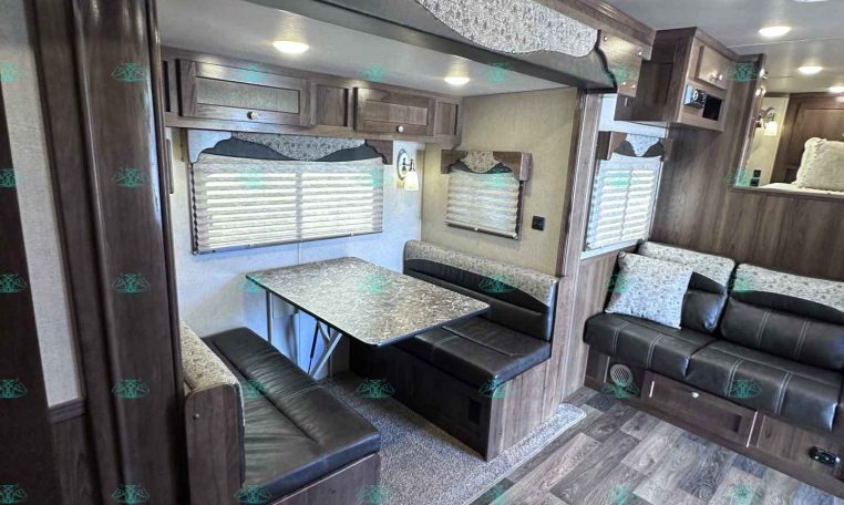 Interior of a recreational vehicle showing a booth-style dinette with a metal tabletop and dark leather seating, wood cabinets, and windows with blinds.