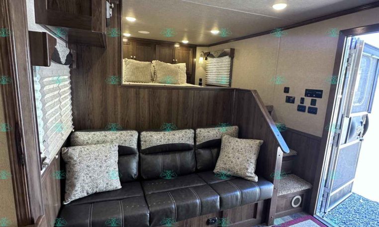 Interior of a wood-paneled RV living area with a black leather sofa and patterned cushions beneath built-in cabinets in warm lighting.