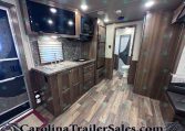 RV kitchen with dark wood cabinets, marble counters, double sink, stove, microwave, and a wall-mounted TV; doorway to the bathroom with toilet nearby.