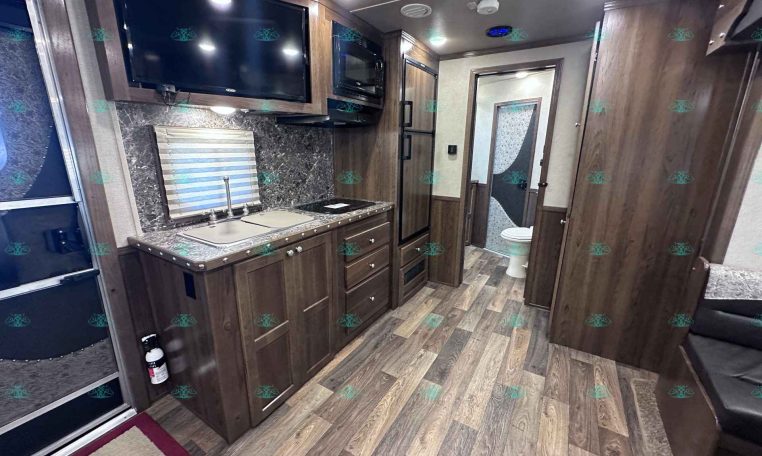RV kitchen with dark wood cabinets, marble counters, double sink, stove, microwave, and a wall-mounted TV; doorway to the bathroom with toilet nearby.