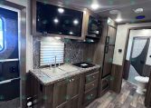 RV kitchen with dark wood cabinets, marble countertop, double sink, stovetop, microwave, and a wall-mounted TV above the counter.