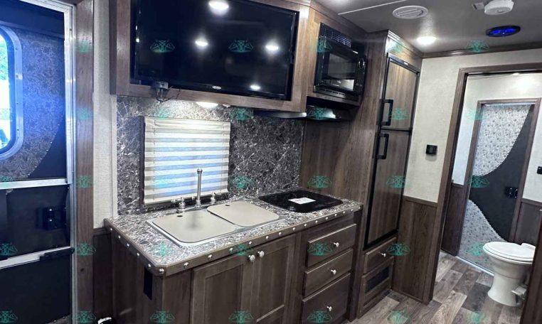 RV kitchen with dark wood cabinets, marble countertop, double sink, stovetop, microwave, and a wall-mounted TV above the counter.