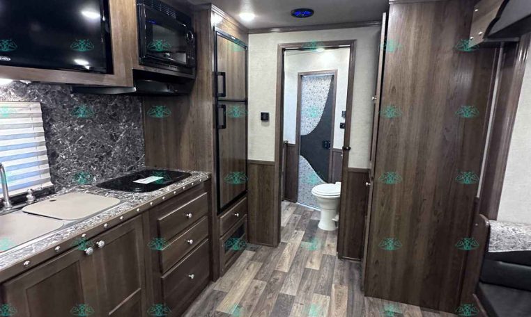 Interior of a modern RV kitchen with dark wood cabinets, marble-style backsplash, microwave, stove, and fridge.