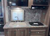 RV kitchen area with dark wood cabinets, marble countertop, and a double sink; a stovetop and microwave are mounted in the upper cabinet, with a large flat-screen TV above the counter and a window with blinds nearby.