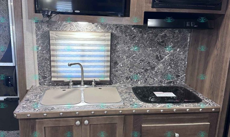 RV kitchen area with dark wood cabinets, marble countertop, and a double sink; a stovetop and microwave are mounted in the upper cabinet, with a large flat-screen TV above the counter and a window with blinds nearby.