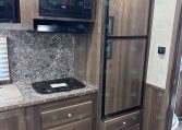 RV kitchen with dark wood cabinets, microwave, stovetop, and tall double refrigerator beside a granite countertop.