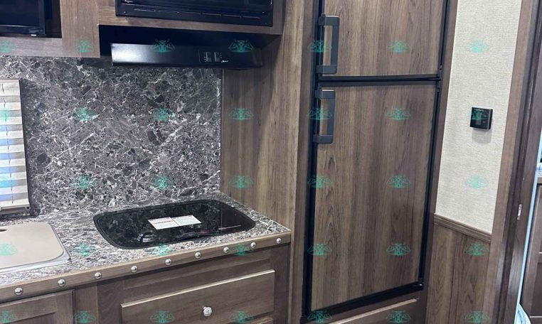RV kitchen with dark wood cabinets, microwave, stovetop, and tall double refrigerator beside a granite countertop.