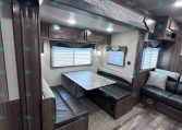 Dark wood RV dining area with a U-shaped booth, aluminum-patterned table, and overhead cabinets; windows with blinds along the side.