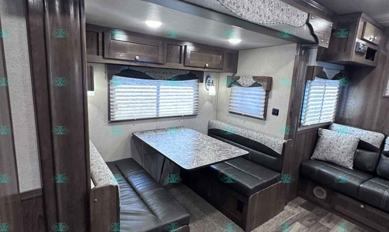 Dark wood RV dining area with a U-shaped booth, aluminum-patterned table, and overhead cabinets; windows with blinds along the side.