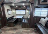 Dining area in a travel trailer: booth seating around a speckled table, with dark wood cabinets and two windows with blinds.