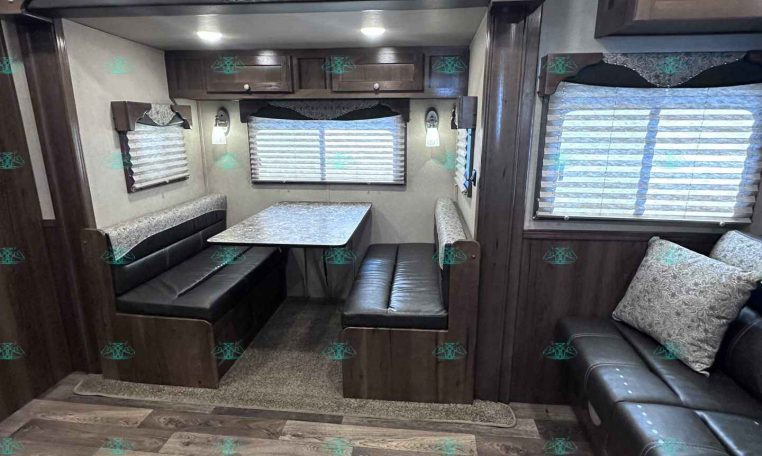 Dining area in a travel trailer: booth seating around a speckled table, with dark wood cabinets and two windows with blinds.