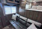 Cozy trailer living area with a dark wood sofa and patterned cushions, bay window with blinds, and built-in cabinets above.