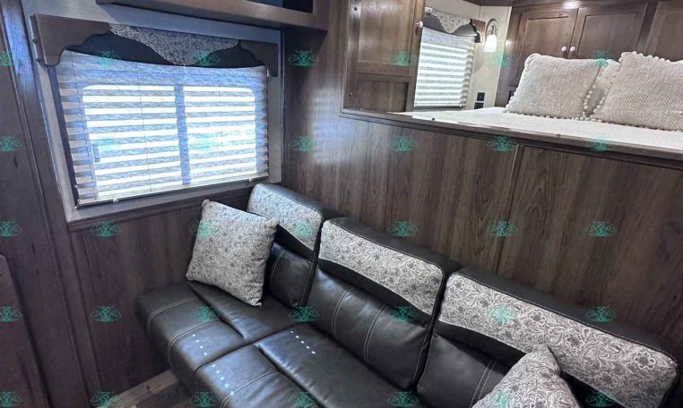 Cozy trailer living area with a dark wood sofa and patterned cushions, bay window with blinds, and built-in cabinets above.
