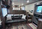 Interior of a wood-toned trailer living area with a black leather sofa and decorative pillows, cabinets above, and a step-up to a bed nook.