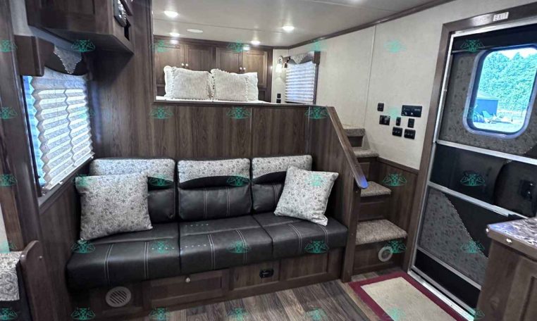 Interior of a wood-toned trailer living area with a black leather sofa and decorative pillows, cabinets above, and a step-up to a bed nook.