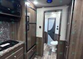 Interior of a modern RV kitchen with dark wood cabinets, stainless appliances, and a marble countertop. A hallway leads to a bathroom at the rear.