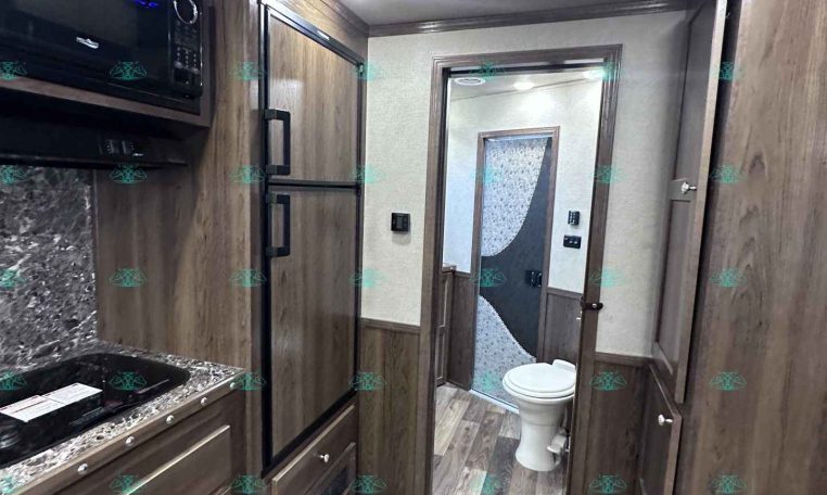Interior of a modern RV kitchen with dark wood cabinets, stainless appliances, and a marble countertop. A hallway leads to a bathroom at the rear.