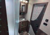 RV bathroom with curved glass shower, dark wood vanity and mirror, small toilet, and textured door panel.